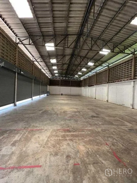 Warehouse for rent in Bang Na, Bangkok - Open-plan warehouse interior featuring a high steel-truss roof, skylights, and concrete flooring with roller shutters along one side.