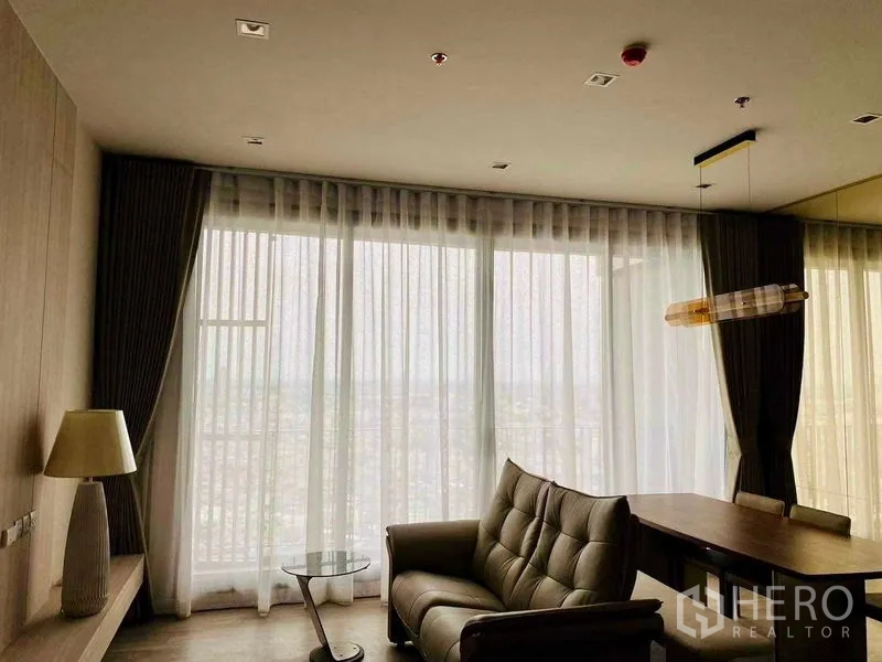 Condominium for rent in Bang Kho Laem, Bangkok - Cozy living room with sofa and desk facing full-height sheer curtains and a broad city skyline view.