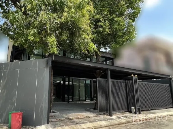Detached House for rent or sale in Phra Khanong, Bangkok - Corner view of gated entrance and large covered carport shaded by a mature tree.