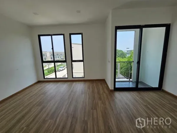 Townhouse for rent in Bang Kapi, Bangkok - Sunny bedroom with wood floors, tall black-frame windows and a sliding door to a small balcony overlooking greenery.