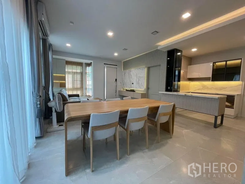 Detached House for rent in Prawet, Bangkok - Bright open‑plan dining and living area with wood dining table, sheer curtains and a modern kitchen backdrop.