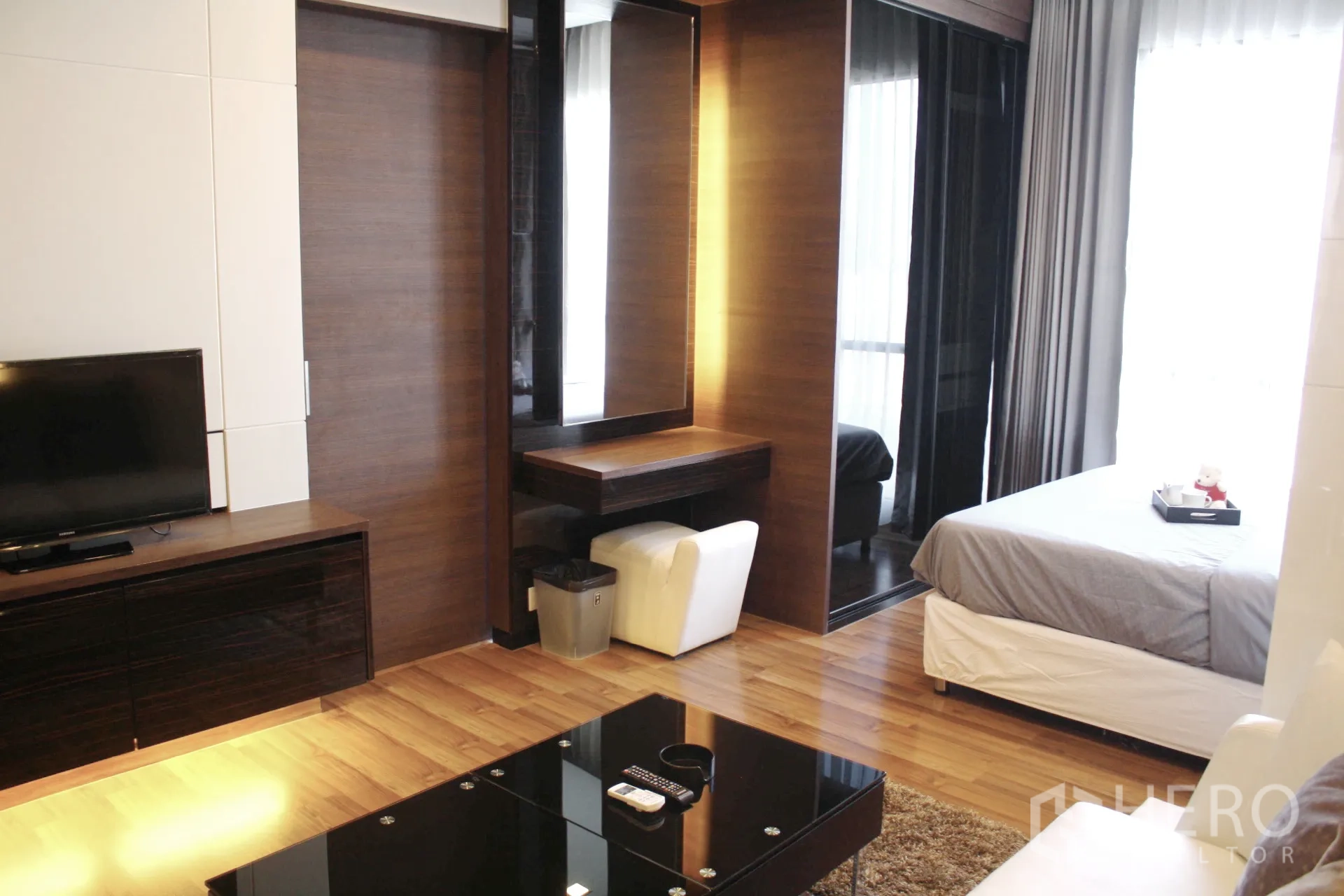 Condominium for rent in Huai Khwang, Bangkok - Open-plan living area with wood floors, TV console, vanity desk, and glass coffee table leading to the sleeping zone.