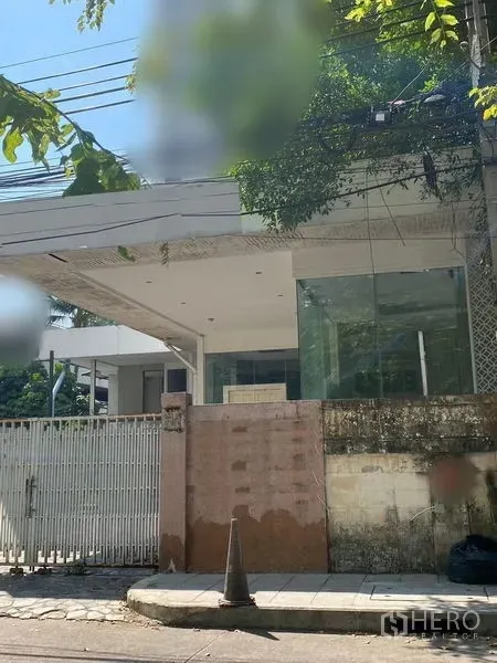 零售空间 出租 - 素坤逸街景中的现代玻璃立面零售建筑，带遮雨门廊、侧边大门、上方电缆与绿荫树木，前方人行道宽阔。