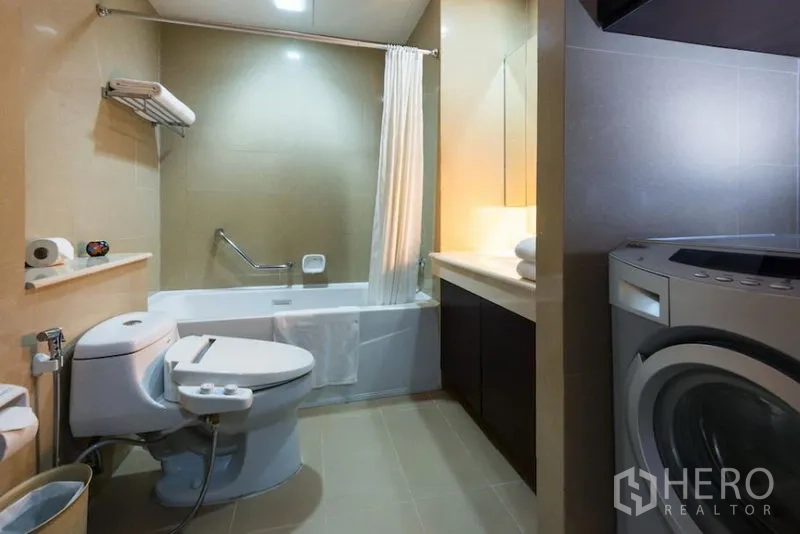 Condominium for rent in Khlong Toei, Bangkok - Bathroom with bathtub, toilet, wide vanity and a washing machine tucked beside the counter.