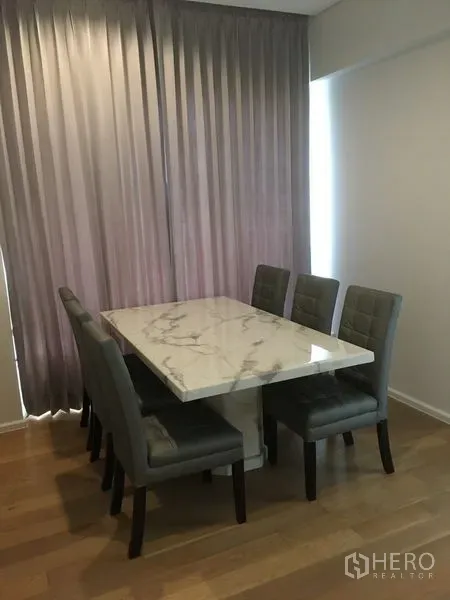 Condominium for rent or sale in Chatuchak, Bangkok - Dining area with a marble-top table and six gray upholstered chairs beside full-height curtains.