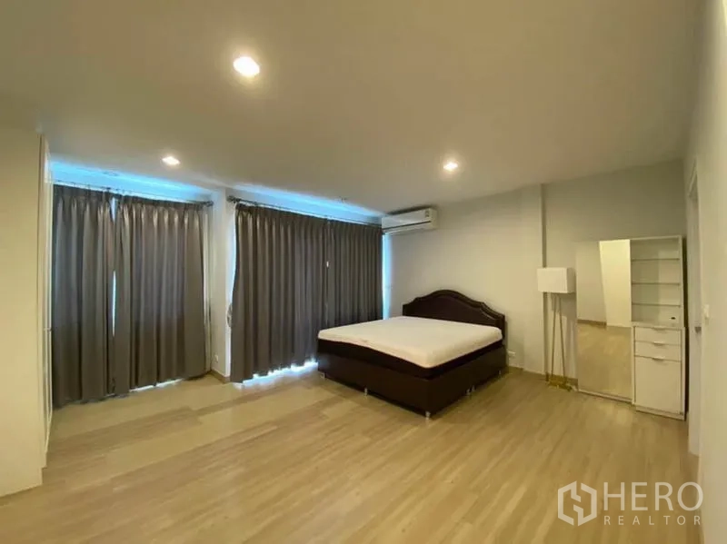 Townhouse for rent in Bang Na, Bangkok - Roomy second bedroom with wood floors, large curtained windows, and air conditioner.