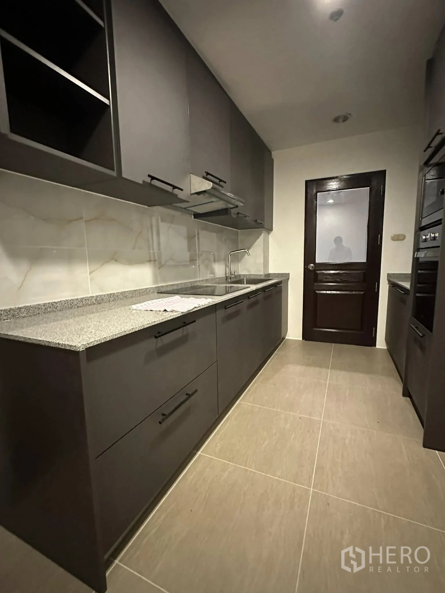 Condominium for rent in Pathum Wan, Bangkok - Galley kitchen with dark cabinetry, stone countertops, and a solid wood door.