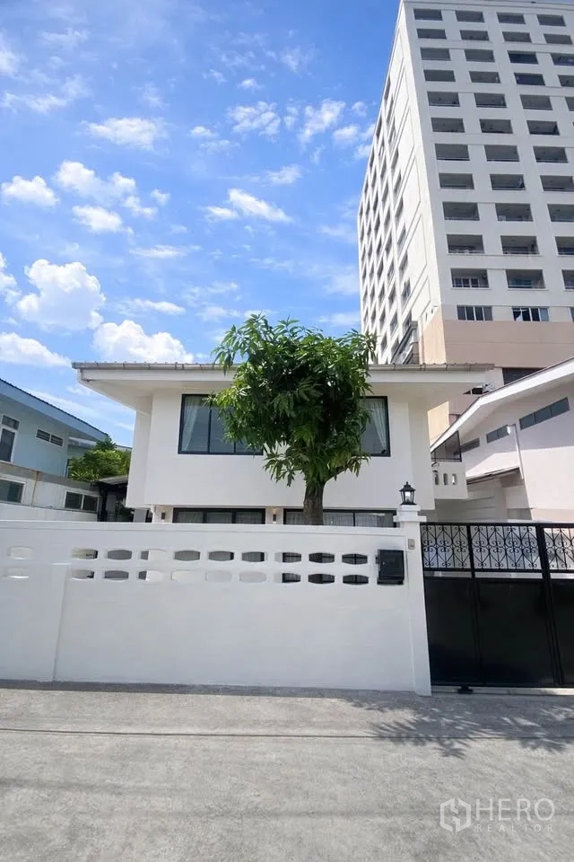 Detached House for rent in Huai Khwang, Bangkok - Street-facing view of the white-walled property with black gate and mature tree.