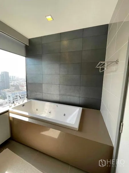 Condominium for rent or sale in Bang Rak, Bangkok - Close-up of the built-in bathtub against a dark tiled wall with a window framing city views.