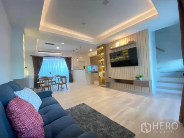 Townhouse for rent in Prawet, Bangkok - Open-plan living and dining area with cove lighting, TV feature wall, wood floors and staircase.
