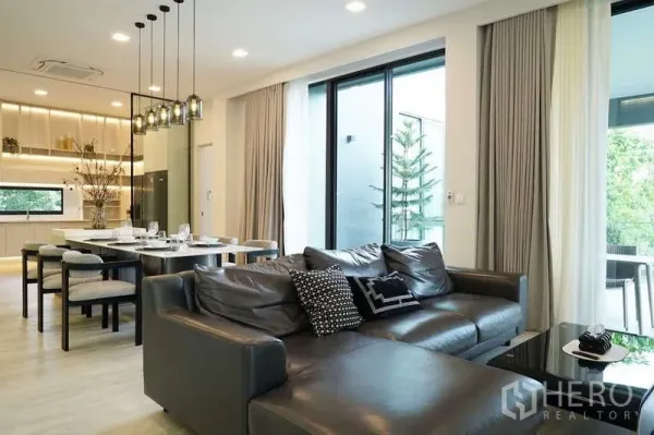 Detached House for rent in Saphan Sung, Bangkok - Open-plan living and dining area with leather sofa, floor-to-ceiling windows and a modern kitchen at Vive Krungthep Kreetha.
