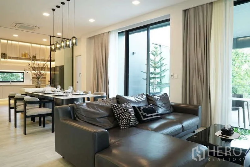 Detached House for rent in Saphan Sung, Bangkok - Open-plan living and dining area with leather sofa, floor-to-ceiling windows and a modern kitchen at Vive Krungthep Kreetha.