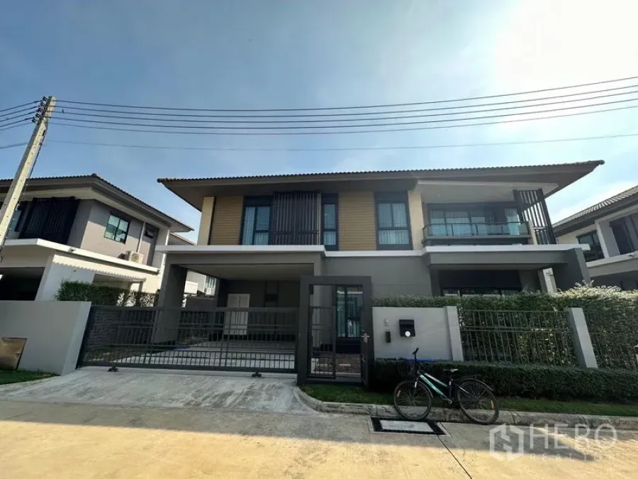 Detached House for rent or sale in Lam Luk Ka, Pathum Thani - Front view of a modern two-storey detached house with a gated driveway and trimmed hedges.