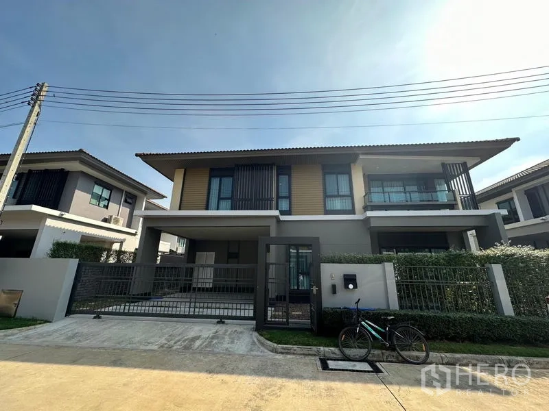 Detached House for rent or sale in Lam Luk Ka, Pathum Thani - Front view of a modern two-storey detached house with a gated driveway and trimmed hedges.