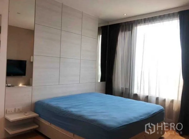Condominium for sale in Khlong Toei, Bangkok - Bright bedroom with large window, sheer curtains and blue upholstered bed.