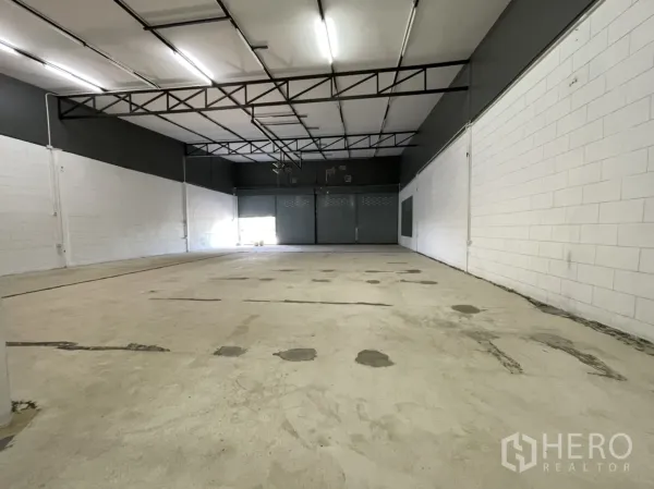 Warehouse for rent in Lam Luk Ka, Pathum Thani - Spacious warehouse interior with high ceiling, steel trusses, bright strip lights and roller shutter doors at the far end.