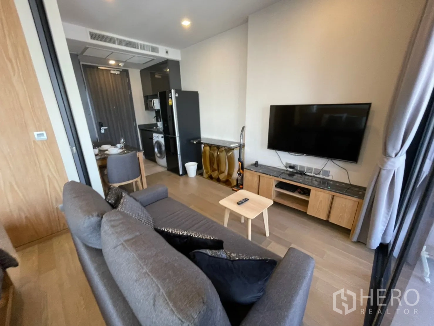 คอนโด for rent in Watthana, Bangkok - พื้นที่นั่งเล่นมีทีวีติดผนัง โซฟาสีเทา และครัวใกล้ประตูทางเข้า