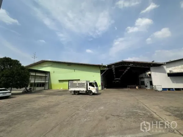 Warehouse for rent in Pak Kret, Nonthaburi - Spacious concrete yard with green and white warehouse buildings, open loading bay and small truck.