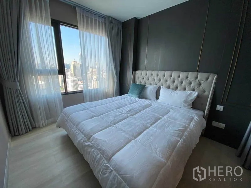 Condominium for rent in Pathum Wan, Bangkok - Bright bedroom with upholstered headboard, sheer curtains and city skyline view.
