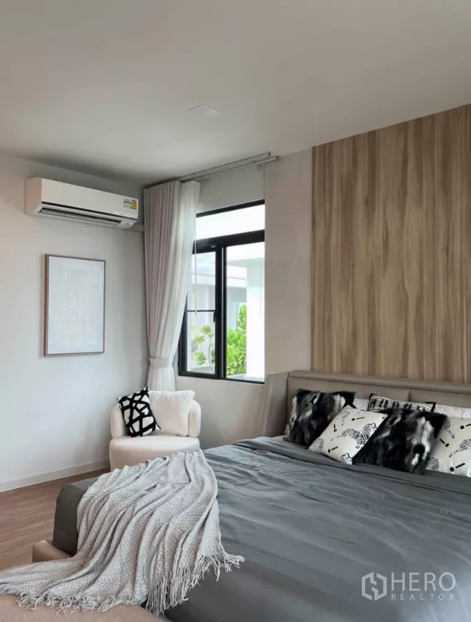 Detached House for rent in Samut Prakan - Bright bedroom corner with AC unit, chair by the window and wood accent wall.