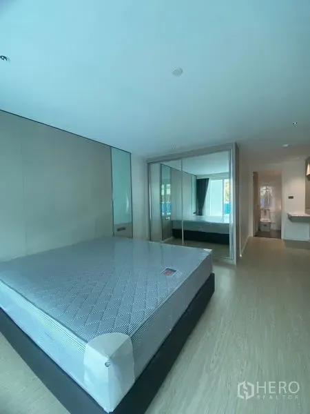 Apartment for rent in Watthana, Bangkok - Primary bedroom with large bed, mirrored wardrobe and doorway to the en‑suite bathroom, finished with light wood flooring.