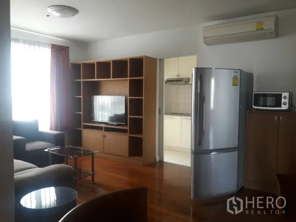 Condominium for rent in Khlong Toei, Bangkok - Cozy living room with TV unit and shelving, sofa set, glass coffee tables and a bright window beside the kitchen.