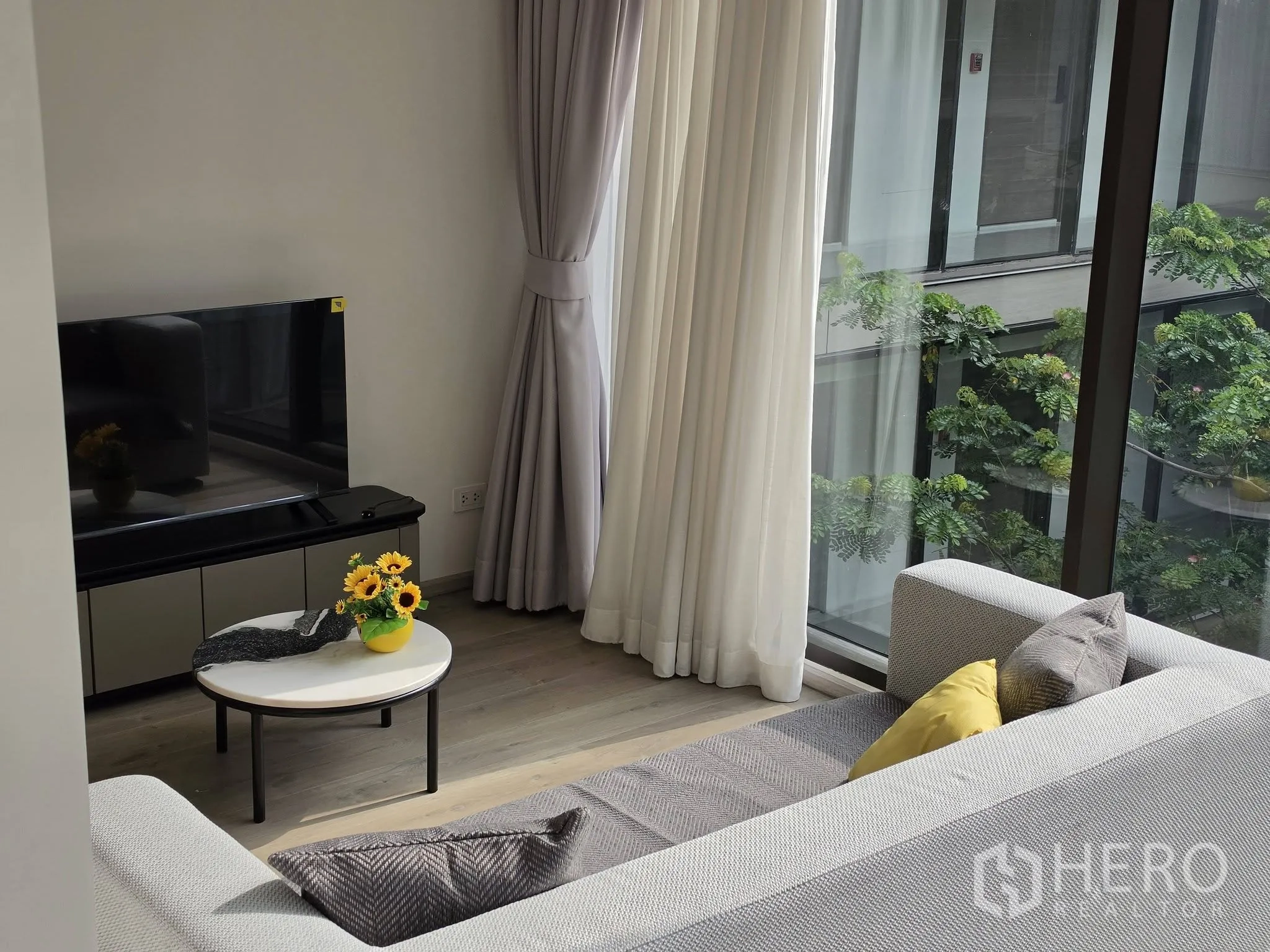 คอนโด for rent in Khlong Toei, Bangkok - ห้องนั่งเล่นสว่างด้วยกระจกสูงจรดเพดาน มีโซฟา ตู้ทีวี โต๊ะกลาง และวิวต้นไม้.