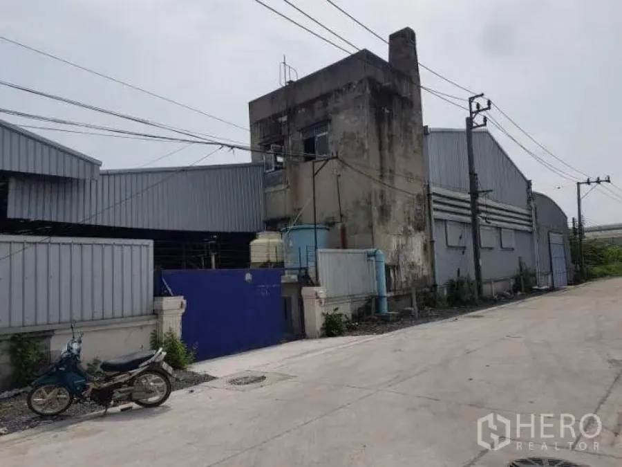 Factory for rent or sale in Bang Phli, Samut Prakan - Gated entry with a blue gate and service building next to linked warehouses on a paved road.