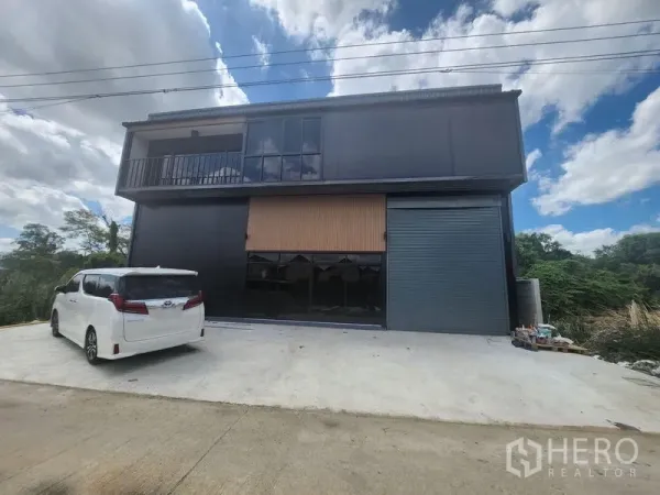 Warehouse for rent or sale in Lam Luk Ka, Pathum Thani - Modern two-storey black warehouse facade with balcony, glass windows and a large roller shutter, plus a concrete driveway for parking.