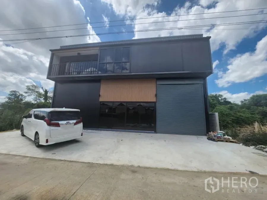 Warehouse for rent or sale in Lam Luk Ka, Pathum Thani - Modern two-storey black warehouse facade with balcony, glass windows and a large roller shutter, plus a concrete driveway for parking.