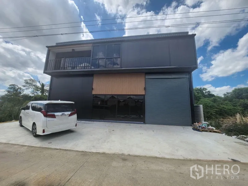 Warehouse for rent or sale in Lam Luk Ka, Pathum Thani - Modern two-storey black warehouse facade with balcony, glass windows and a large roller shutter, plus a concrete driveway for parking.