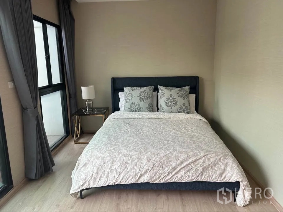 Townhouse for rent in Suan Luang, Bangkok - Cozy bedroom with double bed, side table lamp and corner windows with gray curtains.