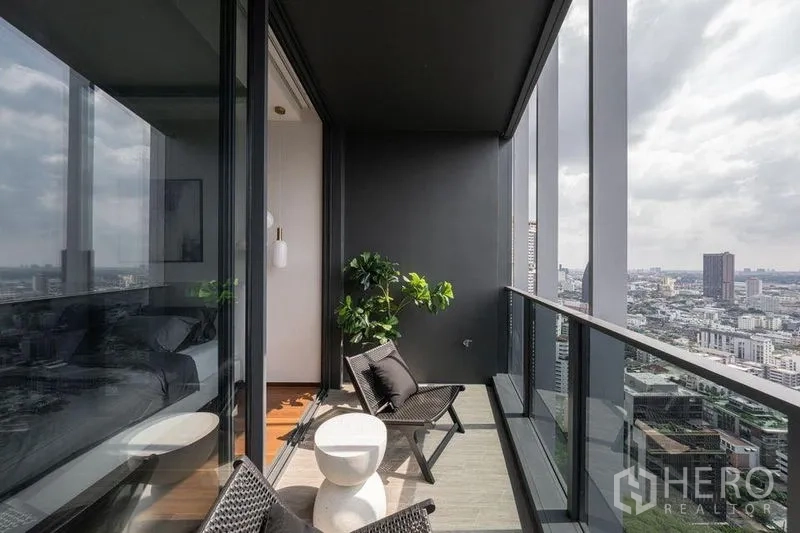 Condominium for rent in Khlong Toei, Bangkok - Covered balcony with lounge chairs, a small table and glass railing overlooking Bangkok city.