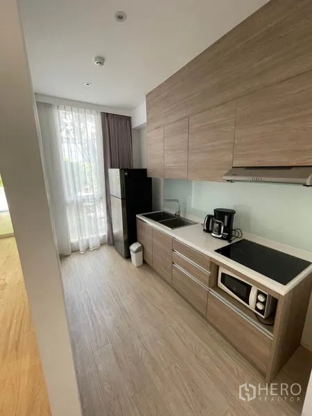 Home Office for rent in Khlong Toei, Bangkok - Compact pantry kitchen with wood cabinets, sink, cooktop, hood, microwave, refrigerator and tall window with sheer curtains.