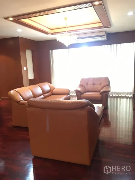 公寓 出租在Watthana, Bangkok - 明亮客厅，浅棕皮沙发，格栅吊顶与水晶吊灯，大窗与木地板。