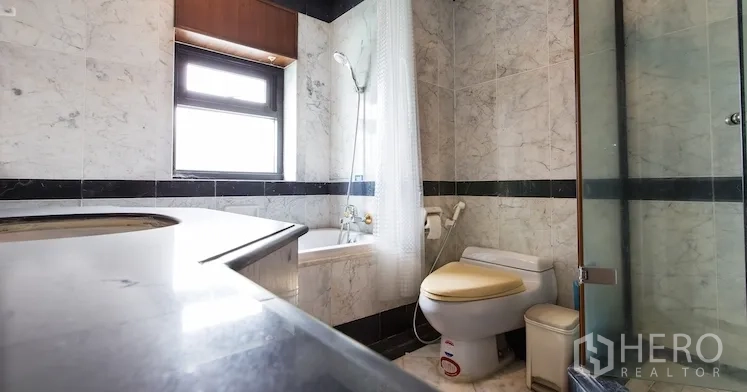 Condominium for rent in Sathon, Bangkok - Marble-tiled bathroom with bathtub-shower combo, toilet and vanity next to a window.