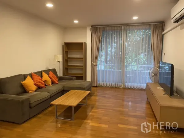 公寓 出租在Pathum Wan, Bangkok - 明亮客厅，L形沙发、木地板、电视柜与落地窗，眺望绿意。