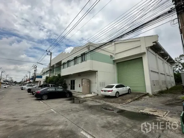 Factory for rent in Khlong Luang, Pathum Thani - Street view of a two-storey factory with a large green roller shutter and on-site parking in an industrial row.