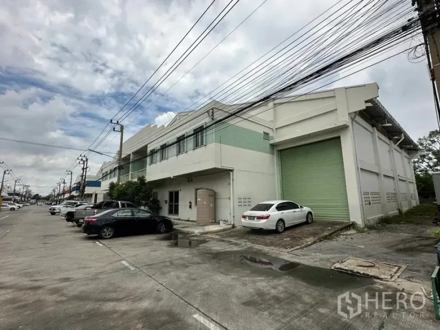 Factory for rent in Khlong Luang, Pathum Thani - Street view of a two-storey factory with a large green roller shutter and on-site parking in an industrial row.