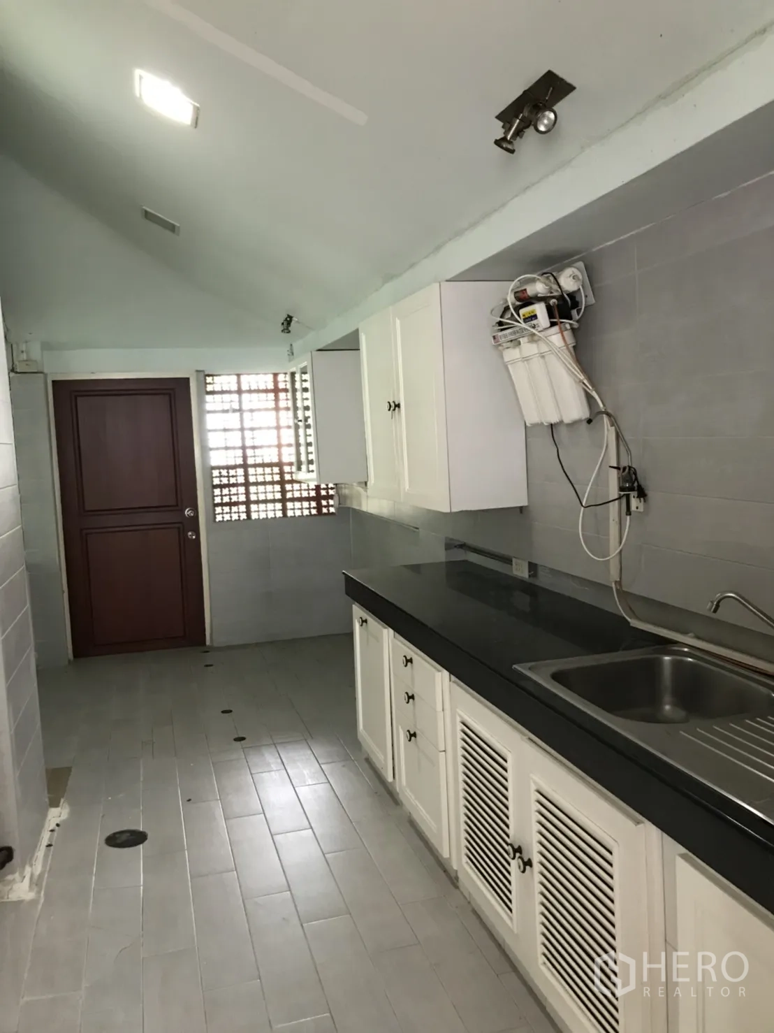 Detached House for rent in Lat Krabang, Bangkok - Galley kitchen with white cabinets, black countertop, stainless sink and grey tile walls.