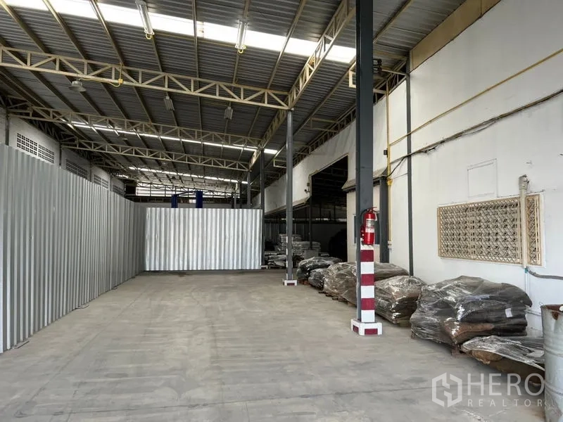 Warehouse for rent in Suan Luang, Bangkok - Open storage bay with concrete floor, corrugated metal partition and fire extinguisher under a lit steel roof.
