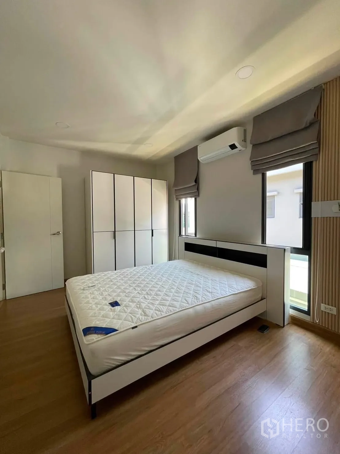 Detached House for rent in Bang Phli, Samut Prakan - Bedroom with white bed frame, wardrobes, air conditioner, two windows with roman blinds, and wood floors.