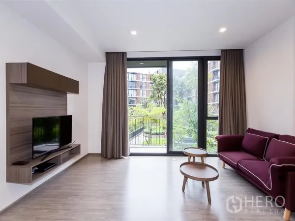 Condominium for rent in Watthana, Bangkok - Bright living room with floor-to-ceiling windows, balcony, TV unit and maroon sofa overlooking a green courtyard at Mori Haus.