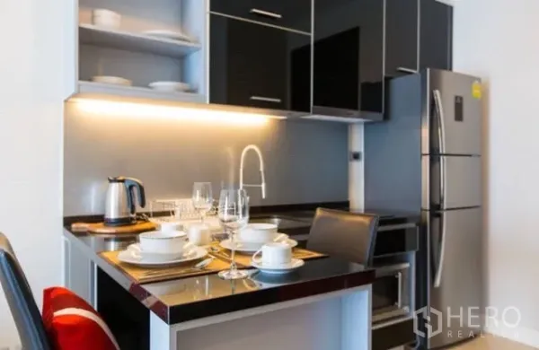Condominium for rent in Khlong Toei, Bangkok - Compact kitchenette with glossy black cabinets, lighting, and stainless fridge.