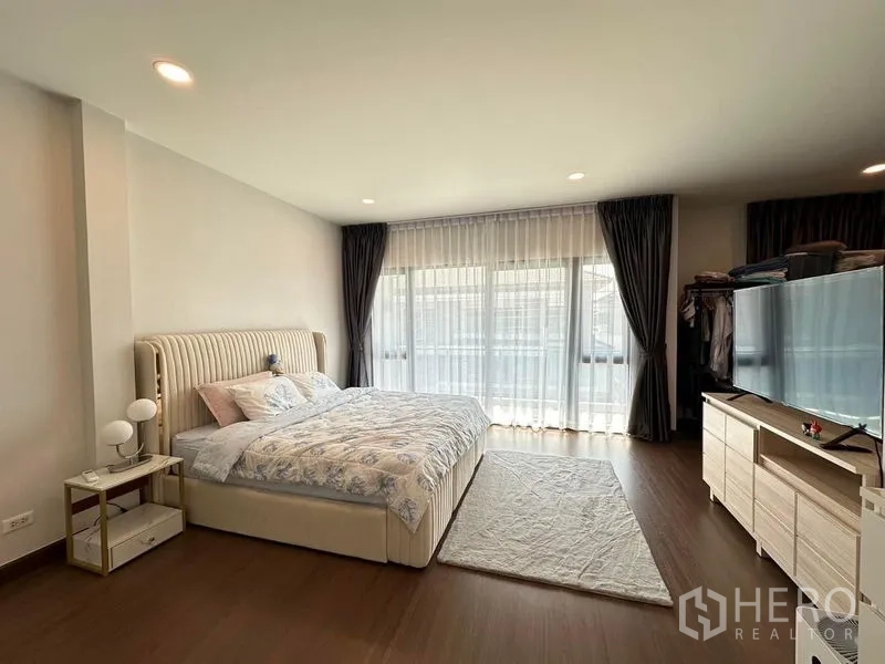 Detached House for rent in Bang Phli, Samut Prakan - Primary bedroom with wall-width windows, king bed, dresser and warm wood flooring.