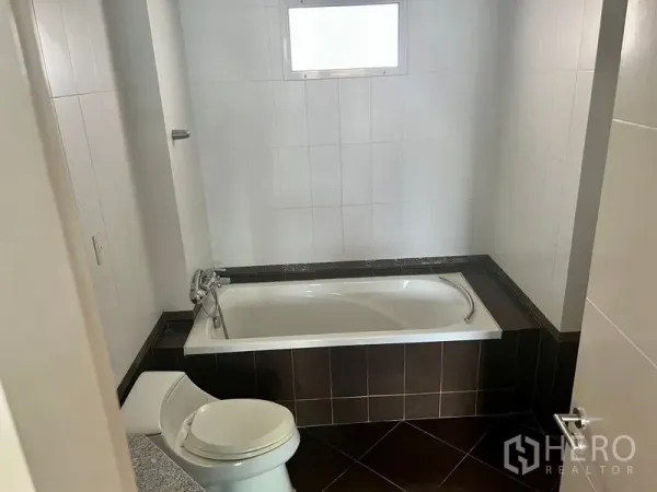 Condominium for rent in Bang Kho Laem, Bangkok - Bathroom with a built-in bathtub, white tiled walls, small frosted window, and dark floor tiles.