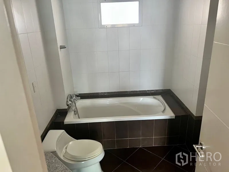 Condominium for rent in Bang Kho Laem, Bangkok - Bathroom with a built-in bathtub, white tiled walls, small frosted window, and dark floor tiles.