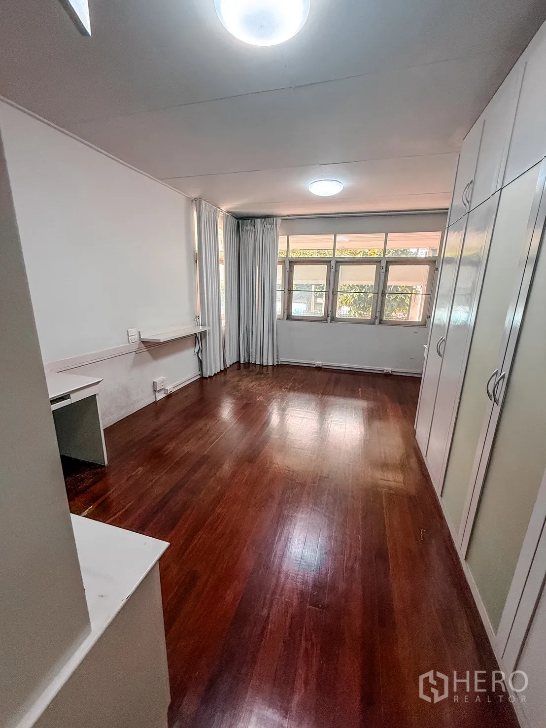 Home Office for rent in Watthana, Bangkok - Long room with hardwood flooring, large curtained windows, built-in wardrobes, and a desk.
