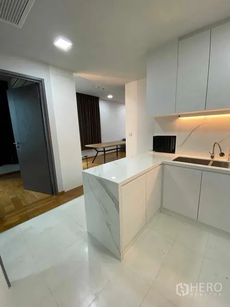 Condominium for rent or sale in Khlong Toei, Bangkok - Kitchen perspective toward dining area showing marble peninsula, sink and bright ceiling lights.