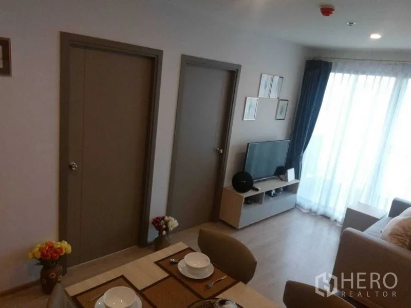 公寓 出租在Bang Na, Bangkok - Bang Na公寓明亮的客餐厅，带电视柜、两扇卧室门和落地阳台窗帘。